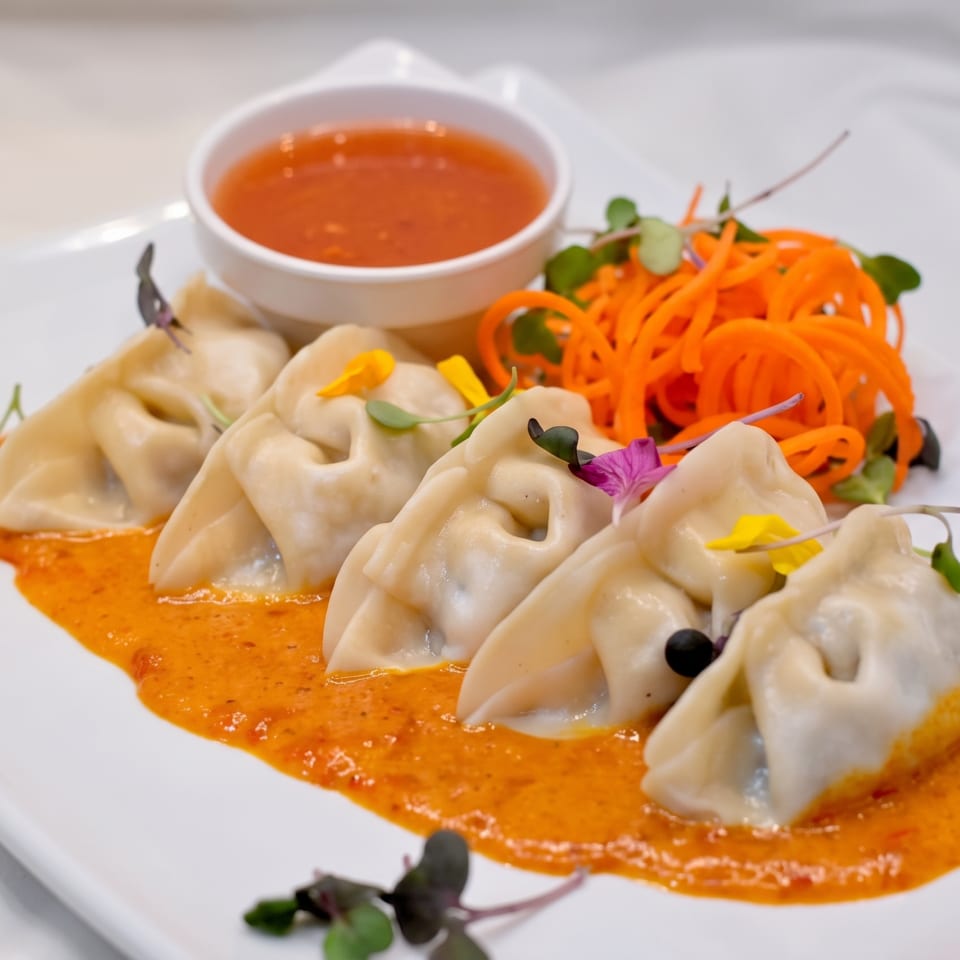 Five dumplings in orange sauce with shredded carrots and a side of red dipping sauce.